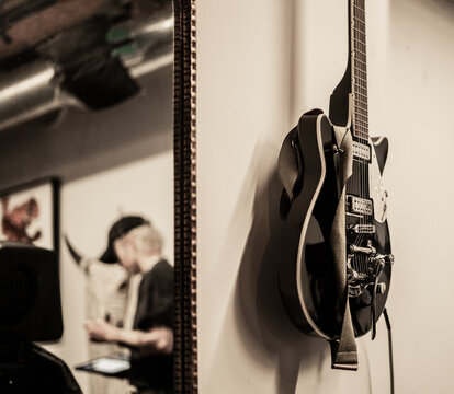 Black electric guitar hanging on a wall inside a music studio with a blurred reflection of a musician