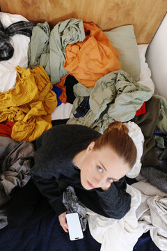 Woman procrastinating laundry in messy bedroom with phone