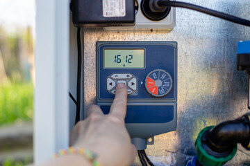A hand operates a digital irrigation timer on a control panel in a garden. smart farming, automated watering, modern technology. © Natalia