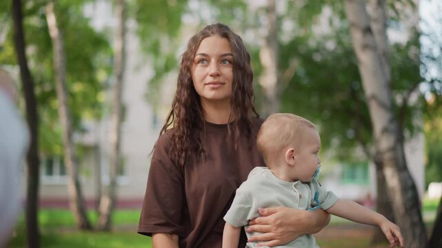 Mother lifting baby in park white woman playful interaction, infant pacifier and curious gaze, birch trees and soft daylight, tender parenting scene with lifestyle decree undertone
