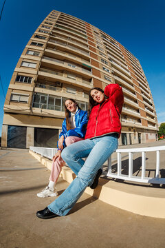Two friends sit on a ledge in front of a tall building in the ci