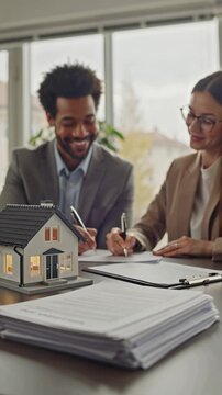 There's a model of a house on the table, and a realtor and a buyer are signing a sales contract nearby.