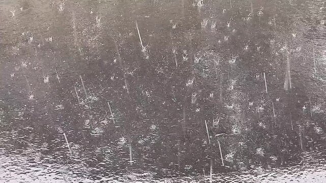 Heavy rain streaks and splashes across a wet asphalt surface, creating droplets, ripples and a moody gray texture. Abstract weather scene ideal for backgrounds, texture overlays and storm concepts