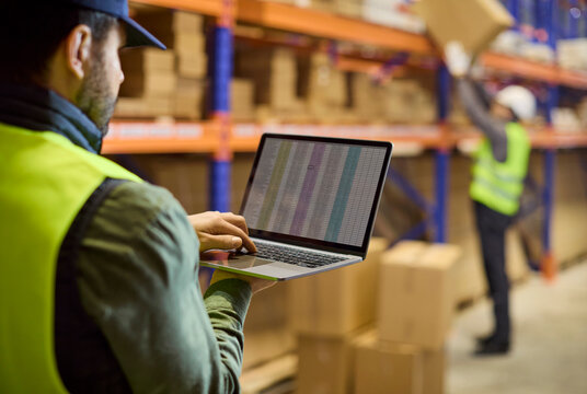 Warehouse inventory worker with laptop near shelves. Staff updates stocktaking data as a colleague moves boxes ensuring storage accuracy and shipping logistics. Clear concept of stock control.