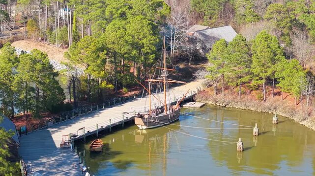 Colonial-era merchant ship replica, the Godspeed, docked at the wooden pier of the Jamestown Settlement living history museum on the James River. Aerial cinematic video.