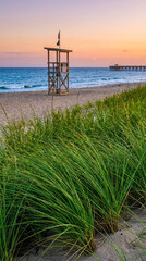 Fototapeta premium Beach scene featuring sea grass, sand, a lifeguard tower, ocean and pier during a colorful sunset, representative of coastal safety and summer tranquility