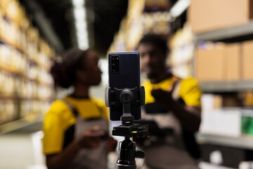 Fototapeta premium Black colleagues filming a marketing vlog in a distribution center, surrounded by cardboard boxes on huge shelves. Team promoting delivery service and e-commerce with live streaming.