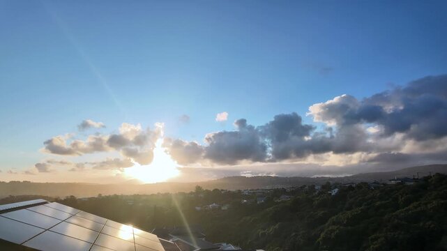 Timelapse of beautiful sunrise over a residential area with solar panels on a house roof. Sunbeams and lens flare illuminating the morning sky