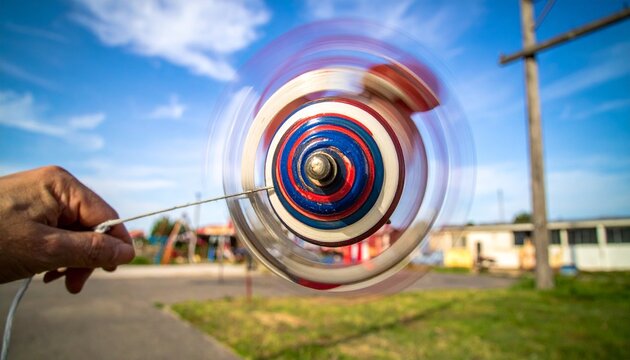A person's hand holding a string, making a colorful spinning top toy whirl and blur against a bright blue sky with clouds
