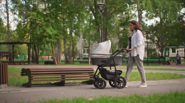 White mother walking stroller in park, sunlit treelined path, wooden bench, gentle pace, checking pram cover, infant inside, calm parenting moment, modern stroller, casual outfit, weekend routine