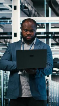 Vertical video Server farm developers using laptop to optimize electronics. Data center teamworking coworkers brainstorming, using notebook device to monitor system performance bottlenecks