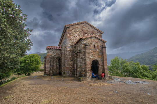 Peque&ntilde;a iglesia rom&aacute;nica de Santa Cristina de Lena hecha de piedra con tejado de teja roja, situada en una colina boscosa bajo un cielo nublado y dram&aacute;tico. Una persona descansa junto a la entrada.