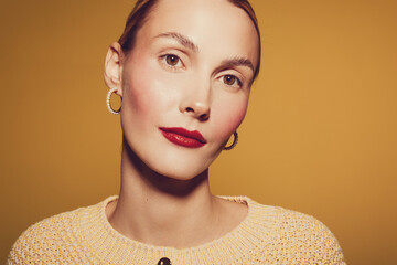 Portrait of a woman with red lipstick and gold hoop earrings wearing a beige sweater. Warm studio lighting highlights her clear skin and calm expression on a yellow background. © SHOTPRIME STUDIO