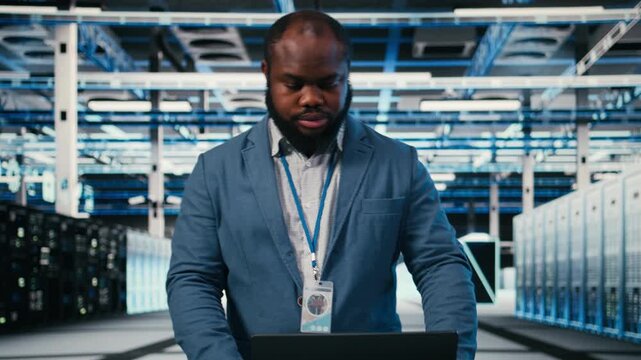 Technician at server room workplace desk using laptop to enhance operational effectiveness of infrastructure. Software developer in data center using notebook to monitor rigs metrics, camera A