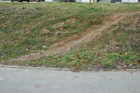 Dirt path leading from grassy slope to paved road, with scattered stones and patches of grass visible along the edge of the pathway