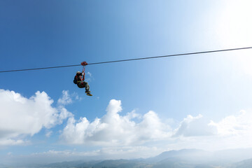 Person zipline gradient blue sky, bright white clouds, blue-gray misty mountains, adventure tourism adrenaline extreme sport freedom courage challenge © Travel Studio Stock