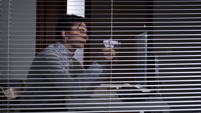 Young man secretly counting stacks of cash behind office blinds, showing concepts of fraud, financial crime, corruption, illegal earnings, and hidden transactions