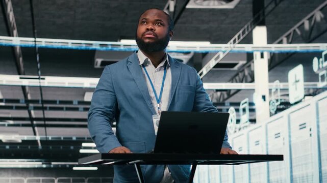 Server farm programmer at workplace desk using laptop to optimize electronics. African american man in data center using notebook to monitor system performance, identifying bottlenecks, camera B
