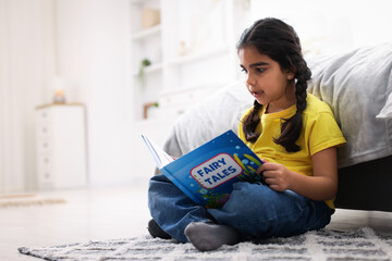 Fototapeta premium Cute little girl reading book on floor at home. Space for text