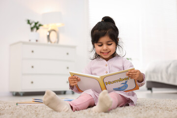 Fototapeta premium Cute little girl reading book on floor at home. Space for text
