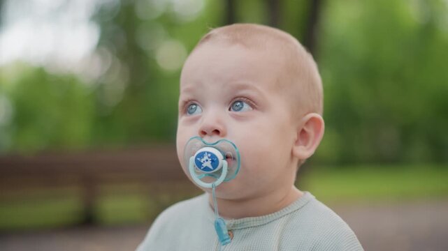 White toddler looking up with pacifier in park, blue eyes fixed on treetop and soft knit sweater visible, bench and green bokeh create calm atmosphere of curiosity, playful routine and family