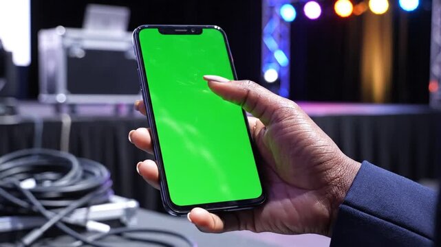 A person's hand holding a smartphone with a green screen, in a stage or event setting with cables and lights.