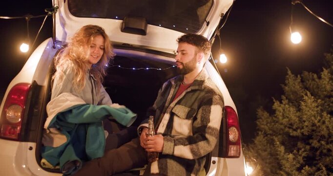 Two people share drinks and talk while sitting in the back of a car under string lights in a camping setting at night with trees around