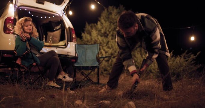 Two friends enjoy time outdoors while cooking and preparing food during a camping trip at night near their parked car