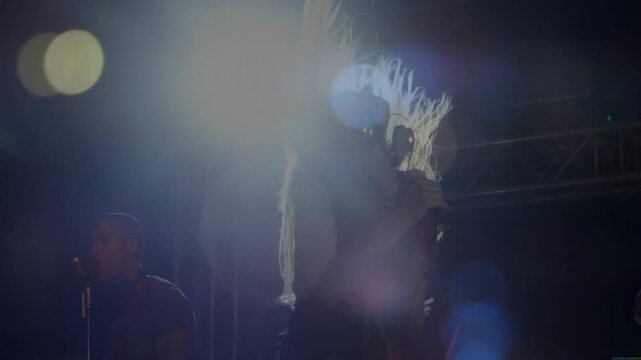 Lead singer onstage grabbing mic moving upright flipping hair backlit lens flares bandmate left