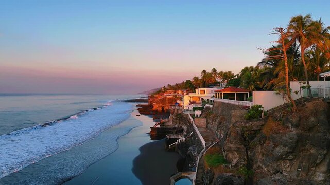 Drone aerial view of sunrise over cliffside oceanfront houses in La Libertad, El Salvador. Pacific waves and luxury coastal living at golden morning light.