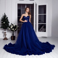 Fototapeta premium A woman poses elegantly indoors, wearing a flowing, deep blue evening gown. The dress cascades to the floor, creating a dramatic effect