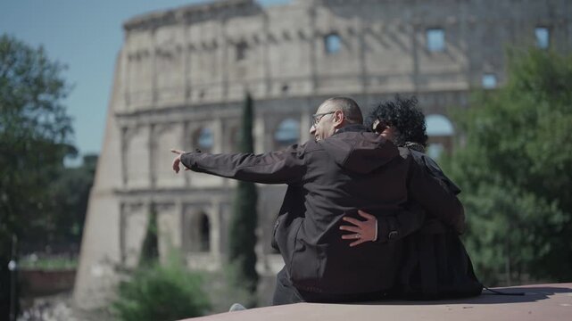 Curly hair woman and men with glasses couple sitting at Parco Celio park look at Coloseum ancient arena and exchange emotions hugging and kissing during sightseeing tour in Rome, Italy, slow motion