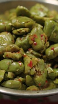 Vertical screen Closeup Pickled Fava Beans In Bowl, Glossy Green Pods Marinated With Chili And Garlic, Vendor Stall Display On Busy Market Counter, Rustic Metal Bowl, Macro Texture, Tangy