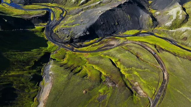 High drone view of a 4x4 car driving switchbacks on a gravel road through a bright mossy volcanic valley with braided river channels in Icelandic Highlands.
