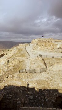 Wide view across Kerak Castle hilltop ruins&mdash;stone ramparts, terraces and courtyards under dramatic clouds with a waving flag, Jordan. Vertical.
