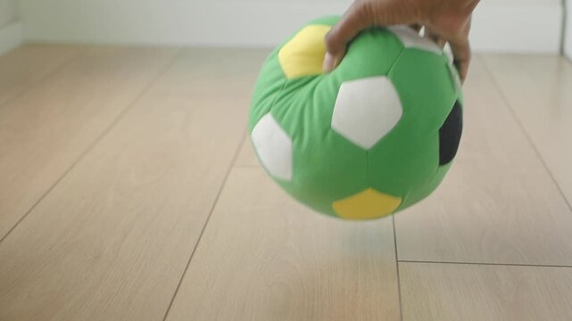 Person picking up green plush soccer ball from wooden floor indoors