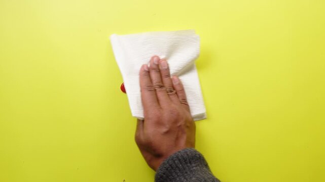 Hands cleaning red tomato ketchup spill from yellow surface with paper napkin