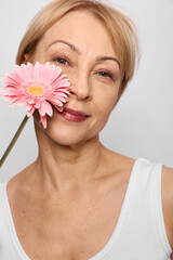Fototapeta premium Mature woman in white tank top holds pink gerbera flower near face, smiling softly with natural makeup on light background. Concept of beauty, aging, wellness, and floral care.