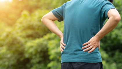 Naklejka premium Person holding aching lower back with hands outdoors with blurred green foliage background