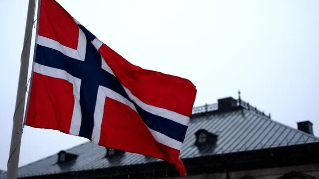 Norwegian flag waving on a pole against a cloudy sky above a building's roof