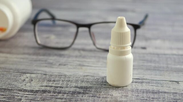 Close up of eye drops bottle and glasses on wooden table with pills falling from bottle