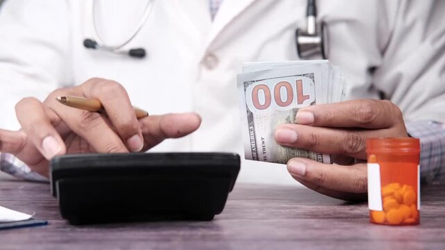 Doctor using calculator while holding stack of hundred dollar bills near pill bottle on desk