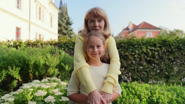 White senior grandmother hugging granddaughter garden, sunny suburban yard with blooming hedge and house, smiling white faces, casual clothes, multigeneration bond between senior people