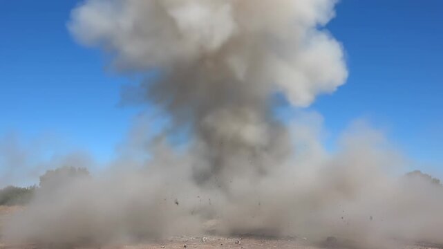 A large gray cloud of smoke rises into a blue sky from a dusty ground