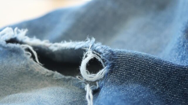 Close up motion of light blue denim fabric with a small hole and frayed threads, showcasing texture and wear in soft focus