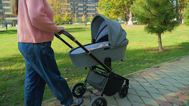 A young woman enjoys a relaxing stroll with her newborn baby in a stroller. Scene set in a city park with green grass and autumn trees on a bright day. Lifestyle concept of parenting and leisure