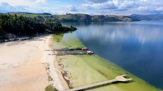 A sunny day at Tota Lagoon, showing wooden piers and red boats on clear water. Green hills surround the scenic landscape.