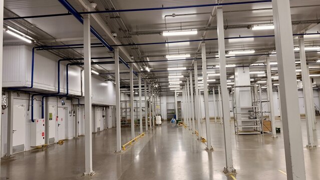 Empty industrial warehouse interior with cooling systems and bright LED lighting. Modern large cold storage facility or factory production hall. Clean floor, metal pillars and white walls.
