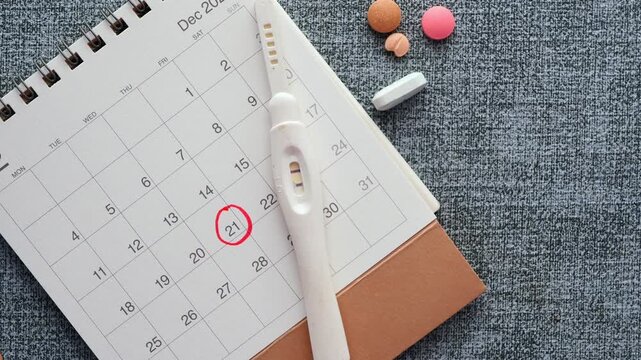 Positive pregnancy test lies next to calendar with marked date, surrounded by various medication pills on gray background, static overhead shot