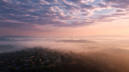 Fototapeta premium Misty dawn landscape with rolling hills and residential areas bathed in soft pastel sunrise light